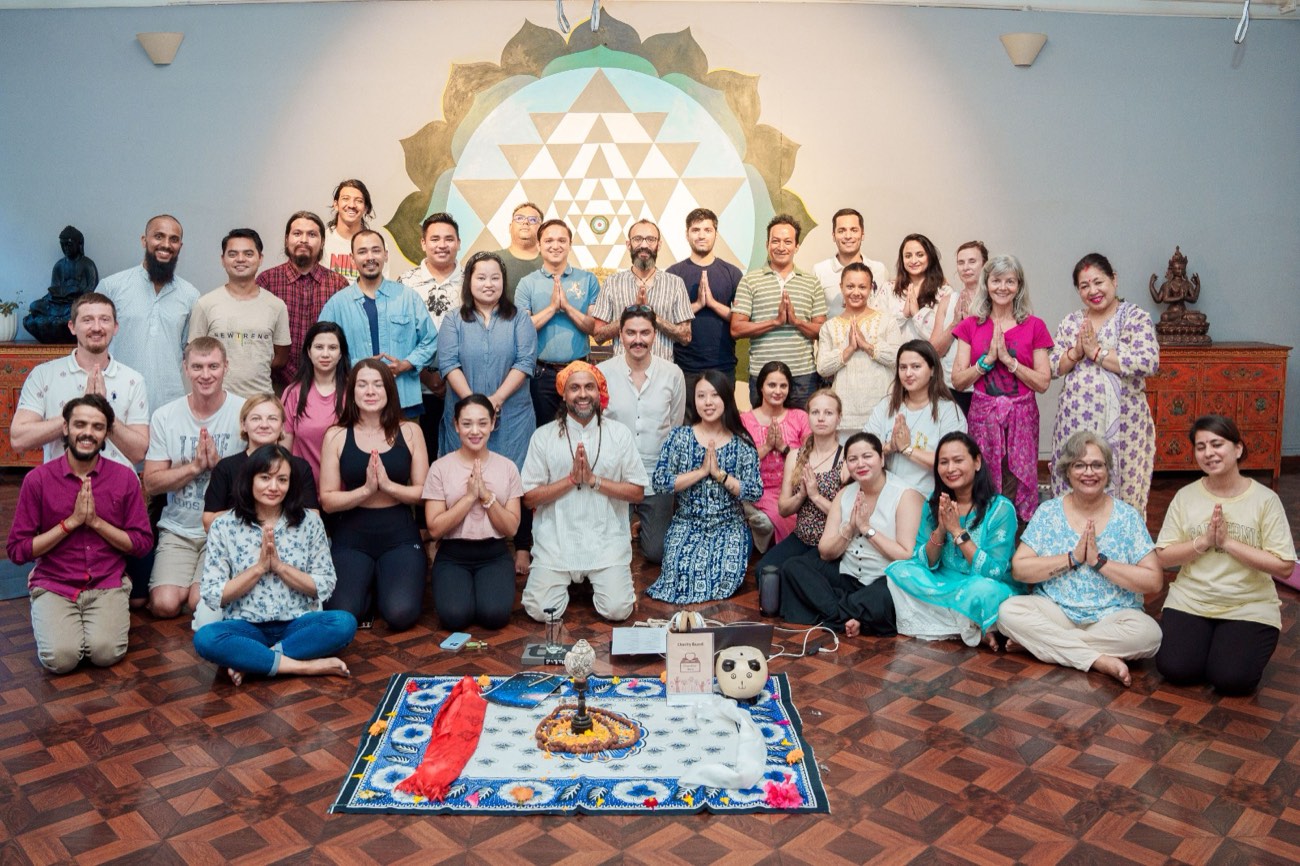 A large group of people with hands in prayer pose after a meditation, in front of a Sri Yantra mural
